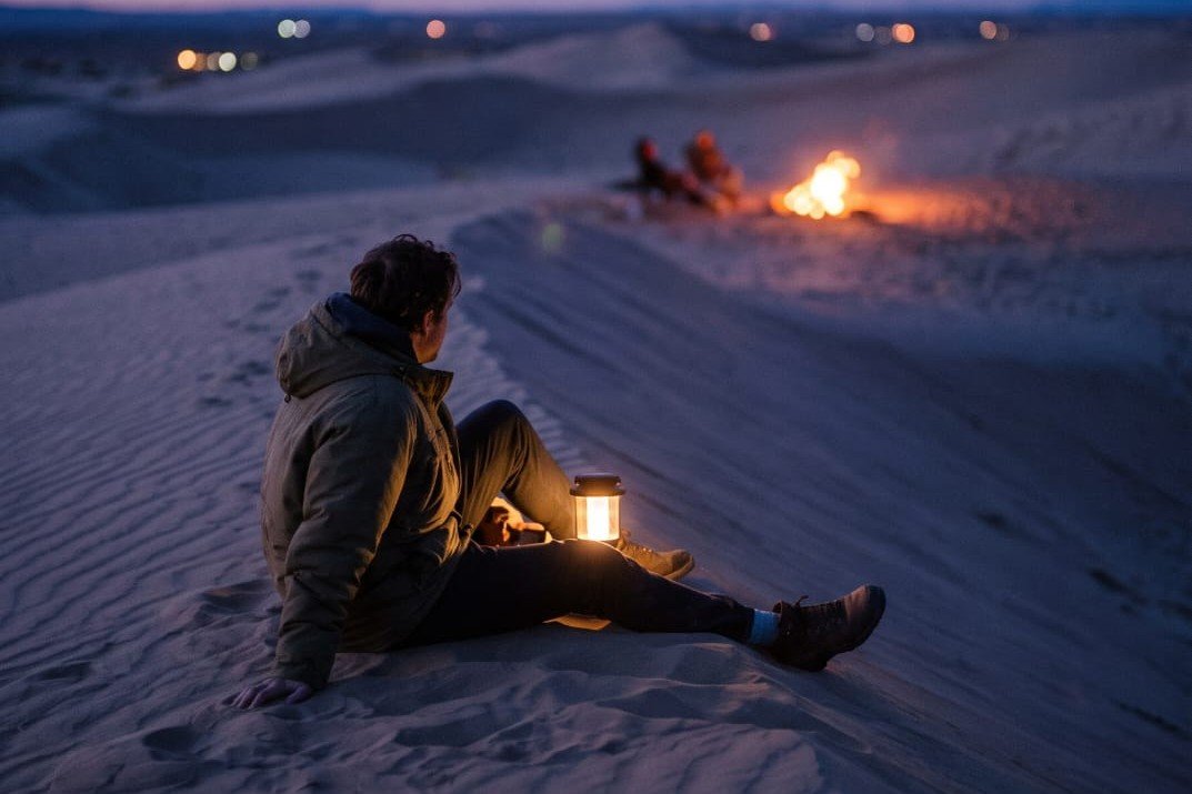 A man on a dune at dusk with a lantern, a fire and two people behind him — House of Bergins