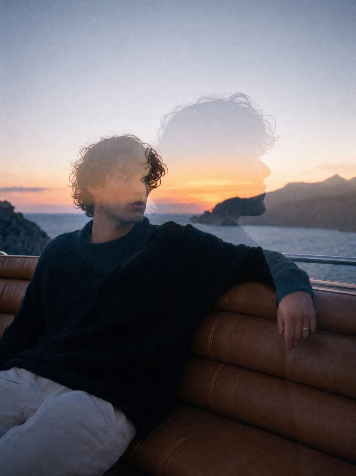 A man on a leather seat at sea at sunset, his profile doubled over the coast — House of Bergins
